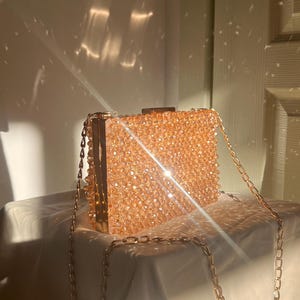 May include: A gold chain purse with a clear, sparkly, peach-colored beaded exterior. The purse is sitting on a white surface.