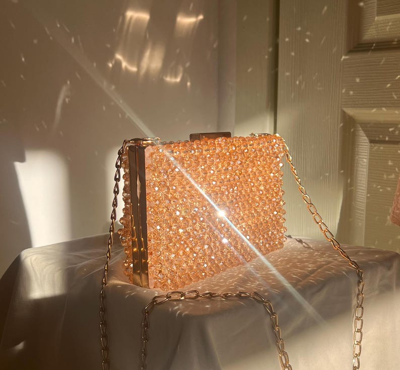 May include: A gold chain purse with a clear, sparkly, peach-colored beaded exterior. The purse is sitting on a white surface.
