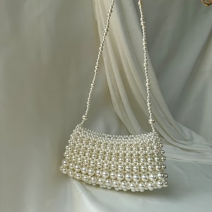May include: A white beaded handbag with a shoulder strap. The rectangular bag is densely covered in small, round, white beads. The strap is made of a strand of smaller white beads. The background is a white fabric.