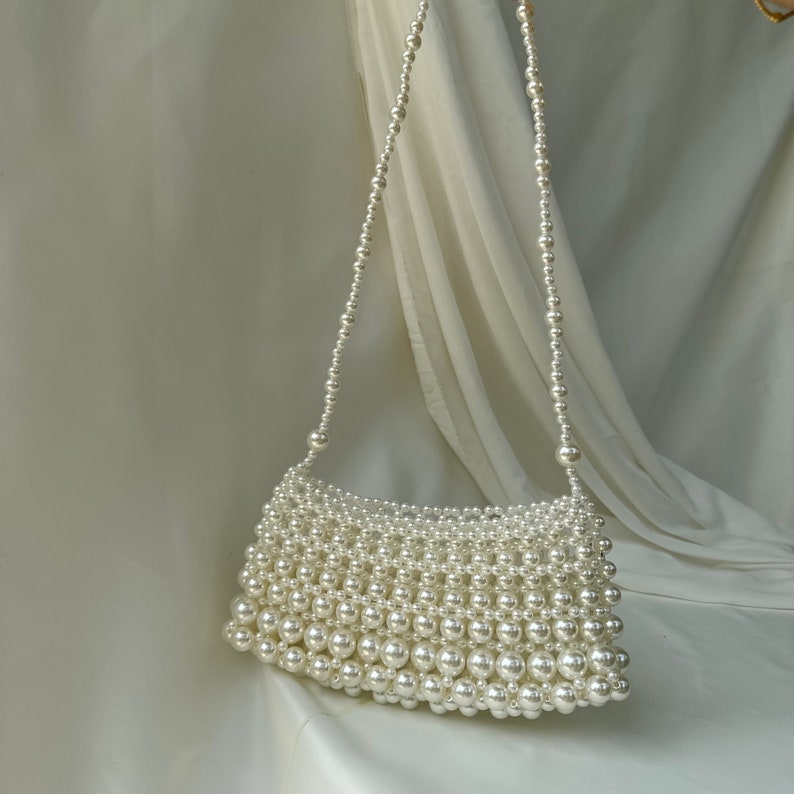 May include: A white beaded handbag with a shoulder strap. The rectangular bag is densely covered in small, round, white beads. The strap is made of a strand of smaller white beads. The background is a white fabric.