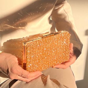 May include: A gold clutch purse covered in small, clear beads. The purse has a gold clasp and a chain strap.