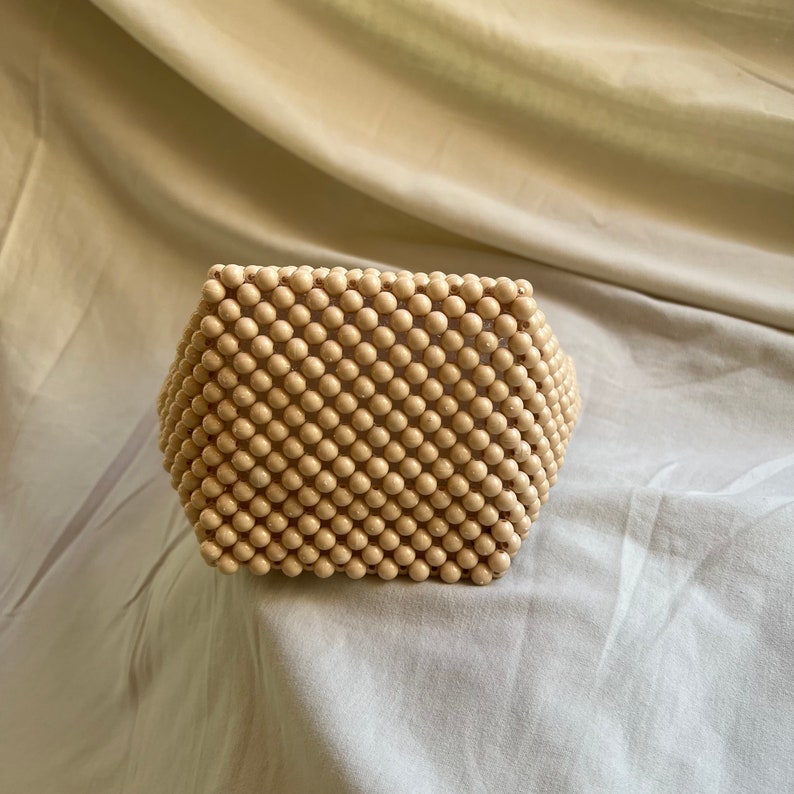 May include: A small, cream-colored beaded bag made of round wooden beads. The bag is square-shaped and has a slightly curved top.