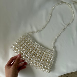 May include: A white pearl beaded handbag with a matching pearl strap. The rectangular bag is made of various sizes of white pearls. The strap is made of smaller pearls. The bag is on a white background.