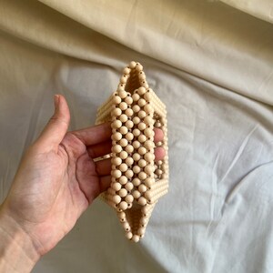 May include: A cream-colored beaded bag with a rectangular shape. The bag is made of small, round beads that are woven together to create a unique and stylish design.