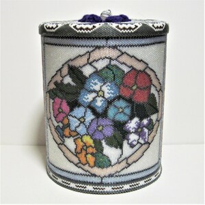 May include: A white cylindrical container with a beaded design and a floral pattern. The container has a purple crocheted lid. The floral pattern features a variety of flowers in shades of pink, blue, purple, red, orange, and green.