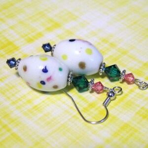 May include: A pair of white, oval-shaped earrings with colorful polka dots. The earrings feature dark blue and green accents, with pink and green crystal beads. The earrings have silver-colored ear wires and are set against a yellow checkered background.