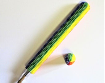 May include: A beaded pen with a matching beaded cap. The pen is covered in small, colorful beads in a pattern of green, yellow, and blue. The cap is a small, round ball with a similar beaded design. The pen is on a white background.