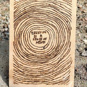 Könnte beinhalten: Ein Holzschild mit einem verbrannten Holzdesign und dem Text "FREEDOM IS A STATE OF MIND" in der Mitte.