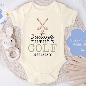 May include: White baby onesie with a brown golf club graphic and the text "Daddy's Future Golf Buddy".