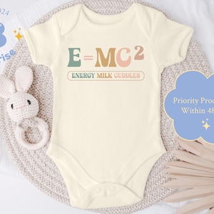 May include: A cream-colored baby onesie with the text "E=MC2" in green, orange, and pink. The text "ENERGY MILK CUDDLES" is written below in a smaller font. The onesie is lying on a beige knitted surface.