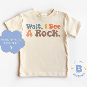 May include: A cream colored t-shirt with the text "Wait, I See A Rock." in colorful bubble letters. The text is in a retro style font. The shirt has short sleeves and a crew neck.
