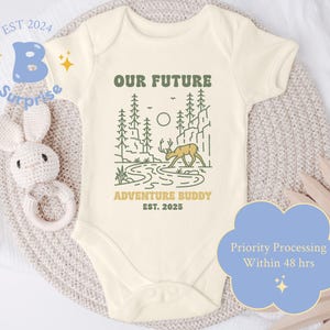 May include: A cream-colored baby onesie with a green and brown illustration of a deer in a forest. The text "Our Future Adventure Buddy Est. 2025" is printed on the onesie.