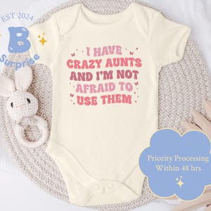 May include: A white baby onesie with pink and red text that reads "I have crazy aunts and I'm not afraid to use them." The onesie is on a light brown knitted surface.