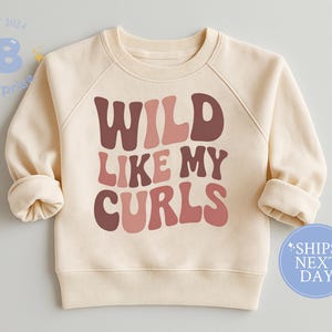 May include: Cream-colored sweatshirt with the text "WILD LIKE MY CURLS" in shades of brown and pink. The sweatshirt has long sleeves and a crew neck. A blue circle with "SHIPS NEXT DAY" is on the bottom right.