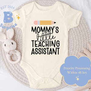 May include: White baby onesie with black text that reads "Mommy's little teaching assistant". A yellow pencil with a pink eraser is above the text.  The onesie is on a light brown surface.