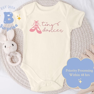 May include: A white baby onesie with the text "tiny dancer" and a pink ballet shoe graphic. The onesie is on a light brown knitted surface.