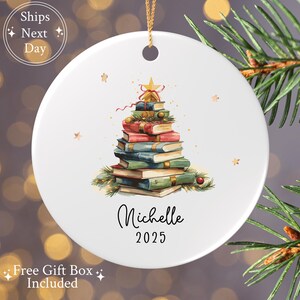 May include: White ceramic Christmas ornament featuring a stack of books arranged as a Christmas tree, adorned with a star and small gold accents. The name "Michelle" and the year "2025" are written in black script.