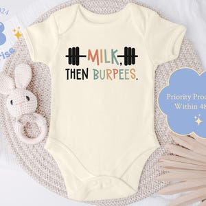 May include: Cream-colored baby onesie with the text "MILK, THEN BURPEES" in black, coral, teal, and tan. A black barbell graphic is above the word "MILK". The onesie is on a neutral-toned surface with a stuffed bunny and a blue cloud-shaped graphic.