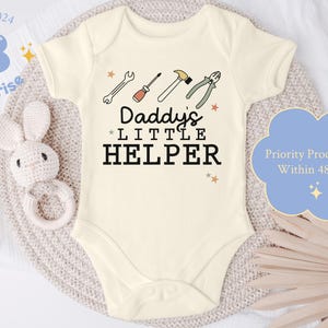 May include: Cream-colored baby onesie with the text "Daddy's Little Helper" and tool illustrations. The design includes a wrench, screwdriver, hammer, and pliers. The onesie is on a neutral-toned surface with a bunny rattle.