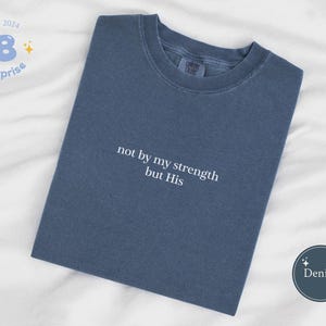 Puede incluir: Una camiseta de mezclilla azul con el texto "not by my strength but His" bordado en blanco. La camiseta está colocada sobre una superficie blanca.
