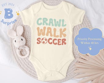 Puede incluir: Un body blanco para bebé con el texto "Crawl Walk Soccer" en una fuente retro con un gráfico de balón de fútbol. El texto está en tonos de verde, naranja y rojo.