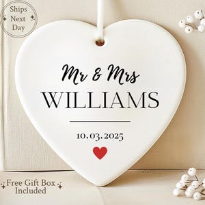May include: A white ceramic heart-shaped ornament with the text "Mr & Mrs WILLIAMS" in black script. Below the names is the date "10.03.2025" and a red heart. The ornament hangs from a white ribbon.