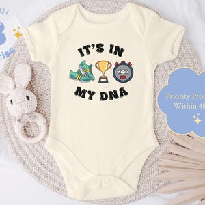 May include: White baby bodysuit with short sleeves featuring a graphic of a trophy, a stopwatch, and a pair of running shoes. The text "It's in my DNA" is printed below the graphic.