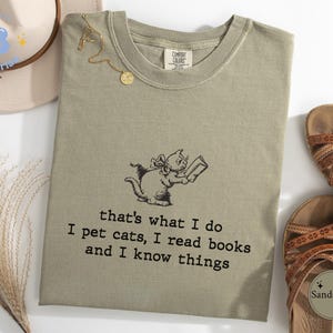 May include: A green t-shirt with a cat reading a book and the text "that's what I do I pet cats, I read books and I know things".