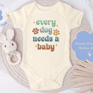 May include: A white baby onesie with a retro-style design that says "every dog needs a baby" in colorful text with paw prints. The onesie is on a light brown textured surface.