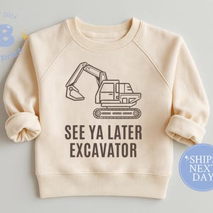 May include: Cream-colored sweatshirt featuring a black excavator graphic and the text "SEE YA LATER EXCAVATOR." The long-sleeved top has a crew neck and rolled cuffs. A blue circle with the text "SHIPS NEXT DAY" is on the bottom right.