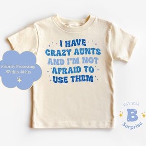 May include: A cream colored t-shirt with blue text that reads "I have crazy aunts and I'm not afraid to use them". The text is in a playful, retro font. The shirt has a crew neck and short sleeves.
