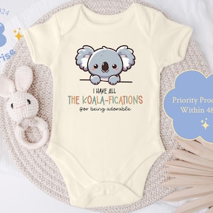 May include: Cream-colored baby onesie with a cartoon koala illustration and the text "I have all the KOALA-FICATIONS for being adorable." A blue cloud-shaped graphic reads "Priority Processing Within 48 hrs."