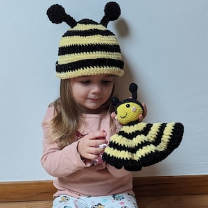 Puede incluir: Una niña pequeña con un gorro de abeja de crochet negro y amarillo sostiene un juguete de abeja de crochet. El juguete de abeja tiene rayas negras y amarillas y una cara sonriente.