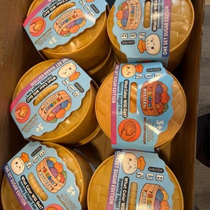 May include: A box filled with several yellow squishy toy sets. Each set includes a round container with a lid and a blue label that reads "BUN Squishy" and "What Color Will Get? Squishy Fidget Fun!" The sets are marked for ages 3+.