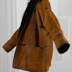 K&ouml;nnte beinhalten: Eine braune Wildlederjacke mit schwarzem Lederbesatz und einem schwarzen Lammfellkragen. Die Jacke hat einen Knopfverschluss und eine aufgesetzte Tasche auf der Vorderseite.