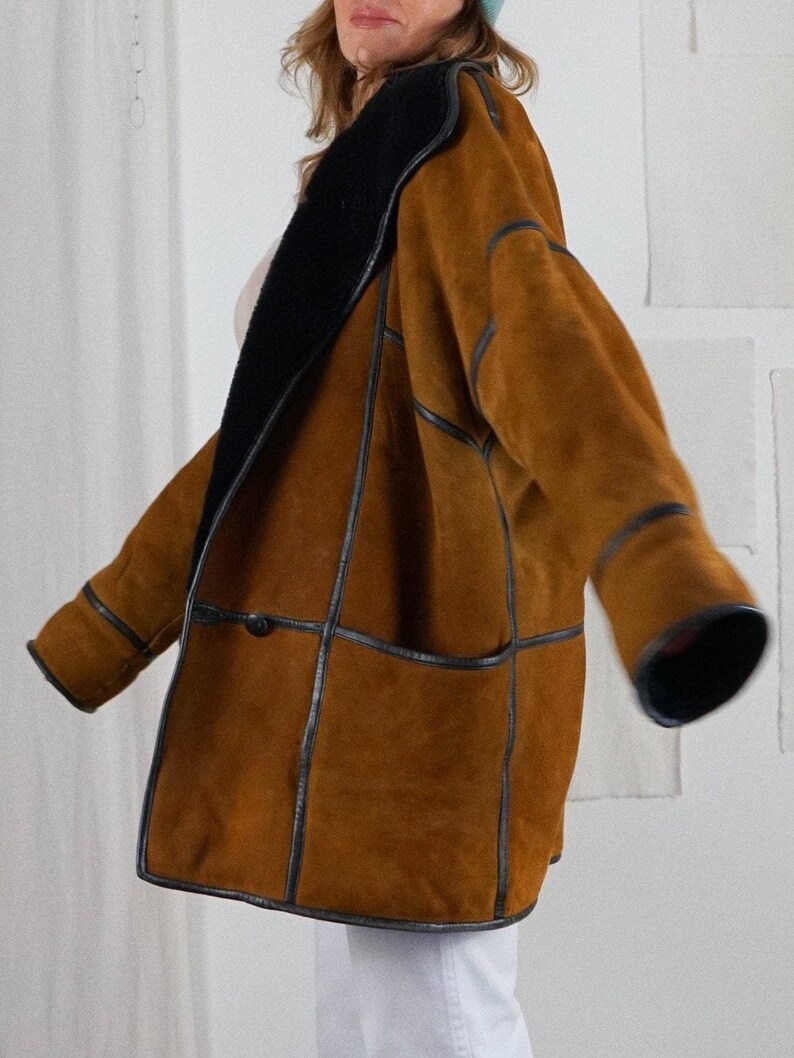 K&ouml;nnte beinhalten: Eine braune Wildlederjacke mit schwarzem Lederbesatz und einem schwarzen Lammfellkragen. Die Jacke hat einen Knopfverschluss und eine aufgesetzte Tasche auf der Vorderseite.