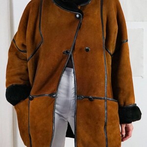 K&ouml;nnte beinhalten: Ein brauner Schaffellmantel mit schwarzem Lederbesatz und einem schwarzen Pelzkragen. Der Mantel hat einen zweireihigen Verschluss mit zwei Kn&ouml;pfen und zwei Taschen.