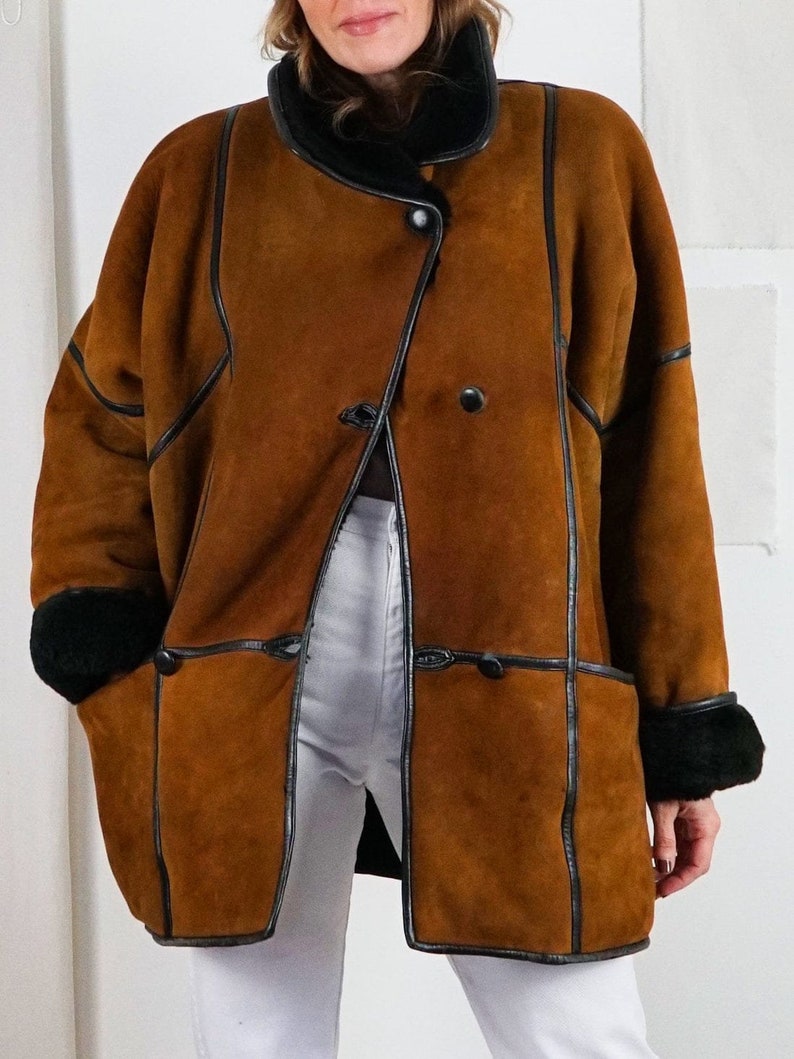 K&ouml;nnte beinhalten: Ein brauner Schaffellmantel mit schwarzem Lederbesatz und einem schwarzen Pelzkragen. Der Mantel hat einen zweireihigen Verschluss mit zwei Kn&ouml;pfen und zwei Taschen.