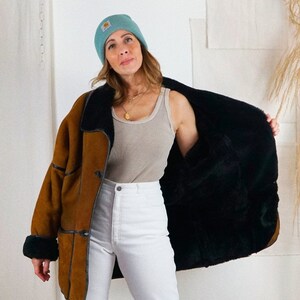 K&ouml;nnte beinhalten: Eine braune Wildlederjacke mit Shearling-Futter und schwarzem Pelz. Die Jacke hat einen Knopfverschluss und einen Kragen. Die Person tr&auml;gt eine wei&szlig;e Jeans und eine hellblaue M&uuml;tze.