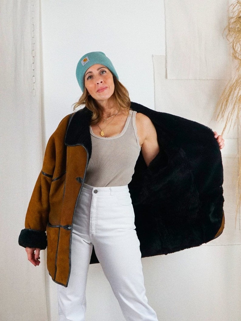 K&ouml;nnte beinhalten: Eine braune Wildlederjacke mit Shearling-Futter und schwarzem Pelz. Die Jacke hat einen Knopfverschluss und einen Kragen. Die Person tr&auml;gt eine wei&szlig;e Jeans und eine hellblaue M&uuml;tze.