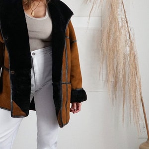 K&ouml;nnte beinhalten: Eine braune Wildlederjacke mit einem schwarzen Lammfellfutter und einem schwarzen Lederbesatz. Die Jacke hat einen Kragen, der hoch oder runter getragen werden kann.