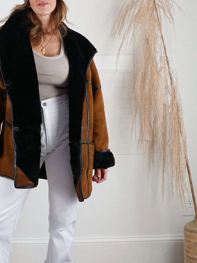 K&ouml;nnte beinhalten: Eine braune Wildlederjacke mit einem schwarzen Lammfellfutter und einem schwarzen Lederbesatz. Die Jacke hat einen Kragen, der hoch oder runter getragen werden kann.