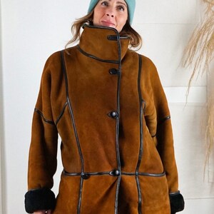 K&ouml;nnte beinhalten: Eine braune Wildleder-Shearling-Jacke mit schwarzem Lederbesatz und einem hohen Kragen. Die Jacke hat einen Knopfverschluss und ist mit schwarzem Fell gef&uuml;ttert.
