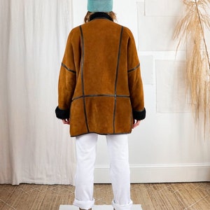 K&ouml;nnte beinhalten: Ein brauner Schaffellmantel mit schwarzem Lederbesatz. Der Mantel wird von einer Person getragen, die eine wei&szlig;e Hose und eine hellblaue M&uuml;tze tr&auml;gt.