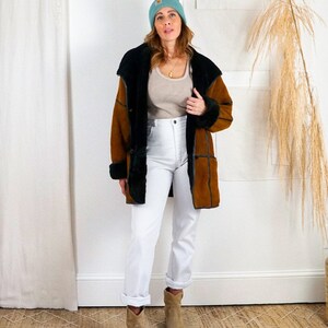 K&ouml;nnte beinhalten: Eine Frau tr&auml;gt eine braune und schwarze Schaffelljacke mit einer hellblauen M&uuml;tze, wei&szlig;e Jeans und braune Cowboystiefel.