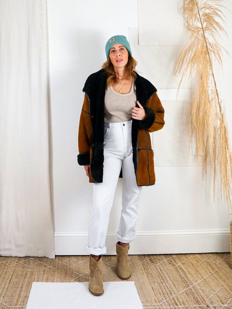K&ouml;nnte beinhalten: Eine Frau tr&auml;gt eine braune und schwarze Schaffelljacke mit einer hellblauen M&uuml;tze, wei&szlig;e Jeans und braune Cowboystiefel.