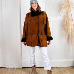 K&ouml;nnte beinhalten: Ein brauner Wildleder-Lammfellmantel mit schwarzem Lederbesatz und einem schwarzen Pelzkragen. Der Mantel hat einen Knopfverschluss und wird &uuml;ber einer wei&szlig;en Jeans und braunen Wildlederstiefeln getragen.