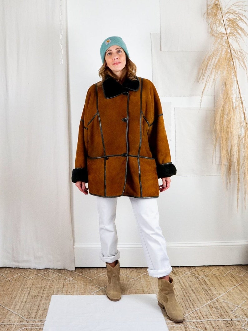 K&ouml;nnte beinhalten: Ein brauner Wildleder-Lammfellmantel mit schwarzem Lederbesatz und einem schwarzen Pelzkragen. Der Mantel hat einen Knopfverschluss und wird &uuml;ber einer wei&szlig;en Jeans und braunen Wildlederstiefeln getragen.