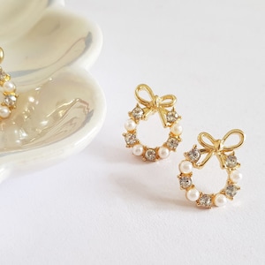 Puede incluir: Un par de pendientes de tono dorado con un diseño de corona. Cada pendiente presenta un pequeño lazo en la parte superior y está adornado con piedras blancas nacaradas.