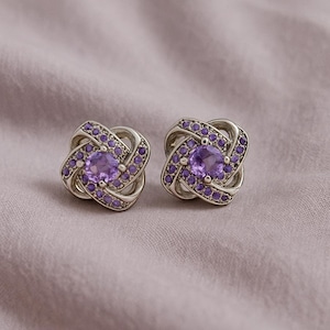 Peut inclure: Deux boucles d'oreilles à clous argentées. Chaque boucle présente une pierre centrale violette, entourée de petites pierres violettes. Le design est en forme de nœud, avec les pierres disposées dans un motif décoratif. Fond en tissu violet clair.